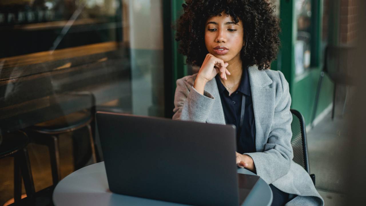 Uma mulher de aproximadamente 24 anos aparece sentada à mesa em frente a um computador.