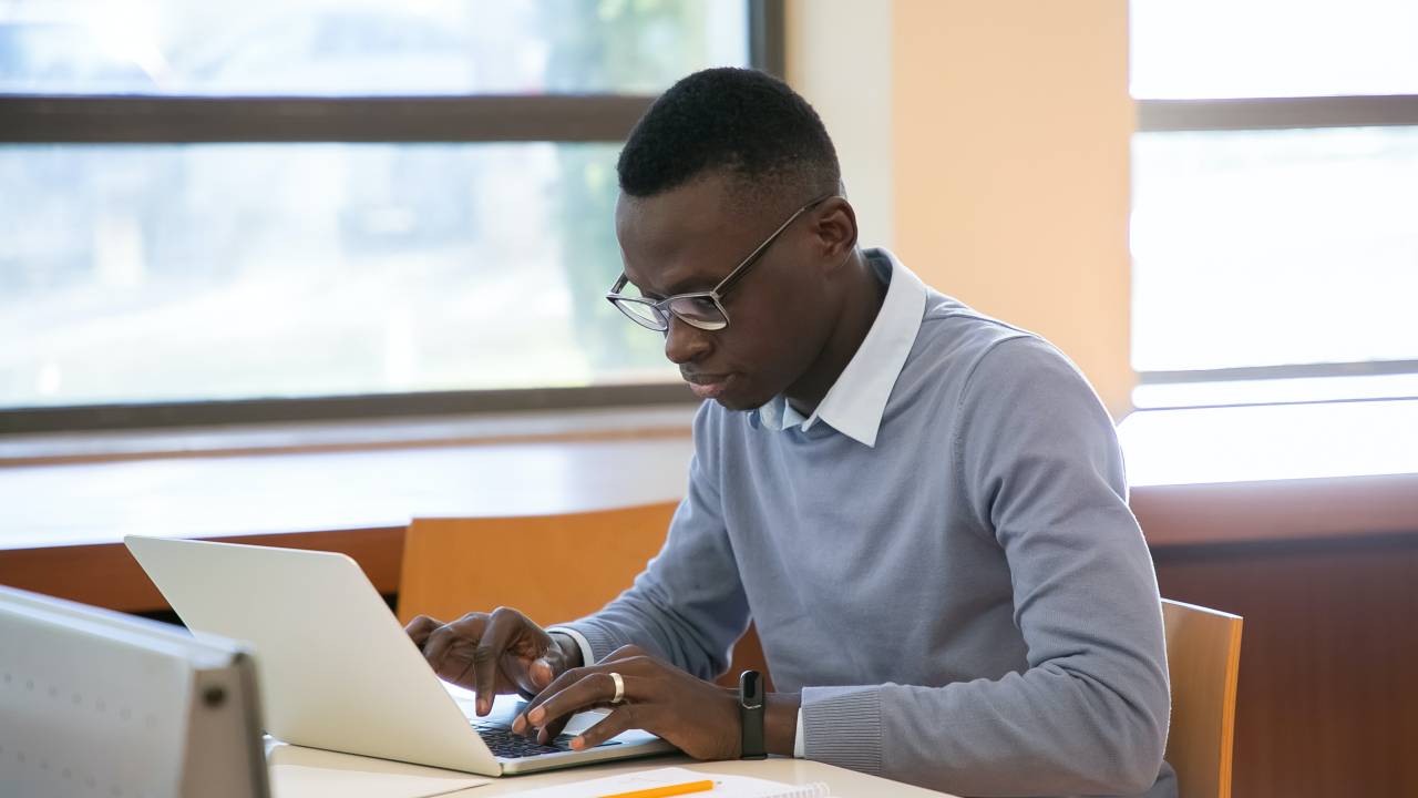 Imagem mostra homem negro com suéter azul usando o notebook em uma mesa ao lado de uma janela.