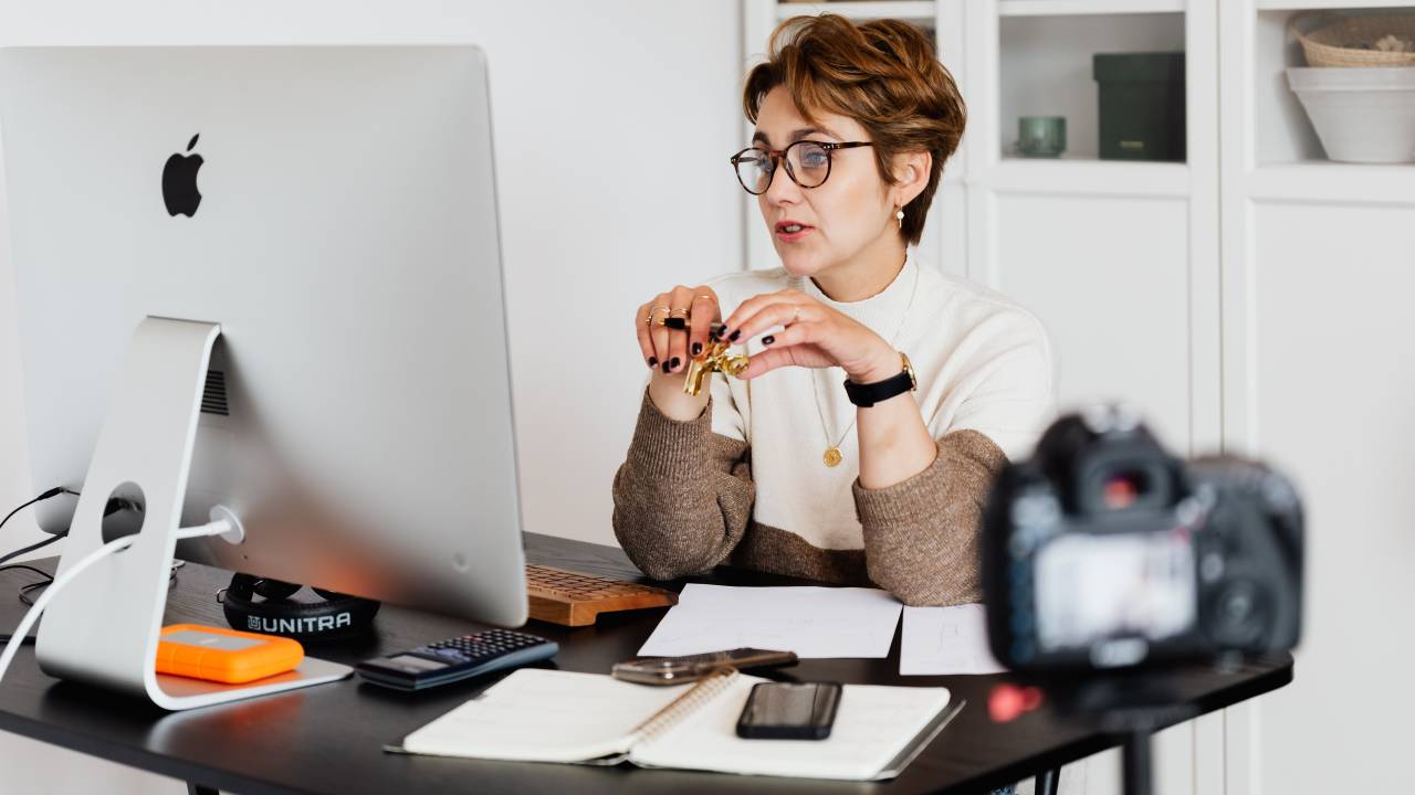 Imagem mostra uma mulher branca, de cabelos curtos, sentada em frente a um monitor, conversando com alguém remotamente.