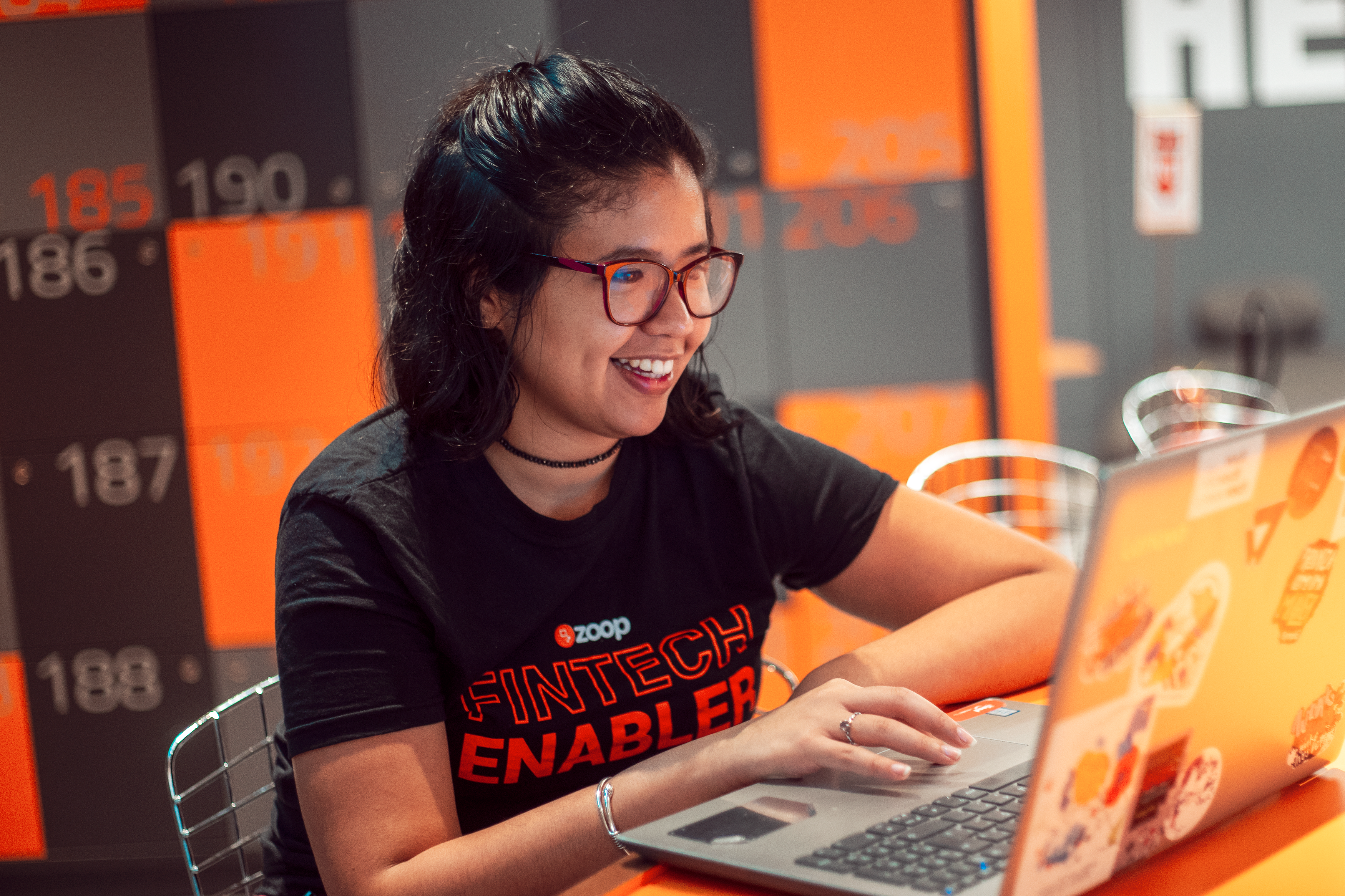 Imagem mostra mulher morena jovem e de óculos, sorrindo ao usar um notebook.