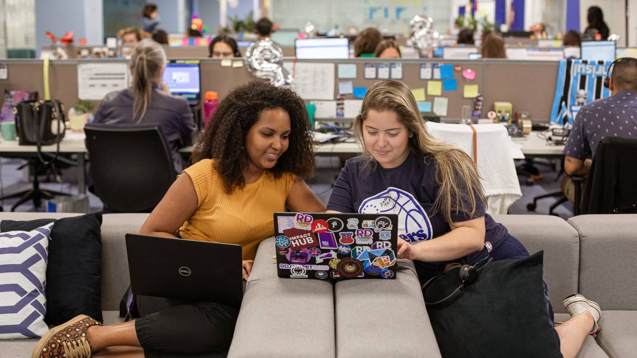 Imagem mostra duas mulheres trabalhando juntas, olhando um notebook. Uma delas é negra de cabelos crespos e escuros e a outra é branca, de cabelos claros e lisos