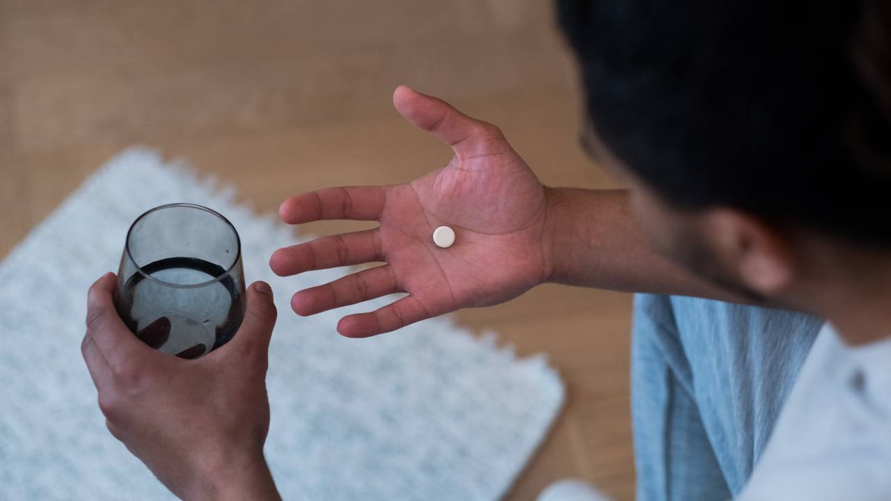 Um homem está sentado olhando para sua própria mão, sobre a qual tem um comprimido. Na outra mão ele segura um copo de água.