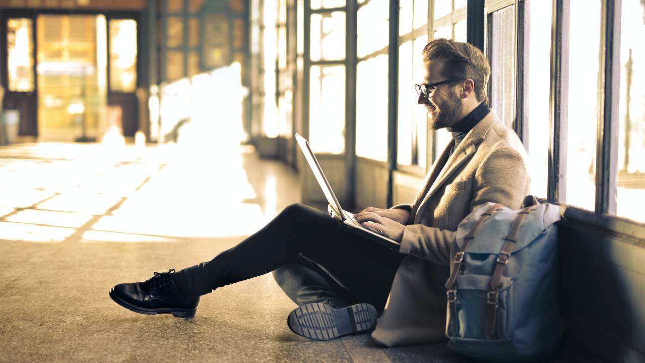 Um homem está sentado em um corredor. Ele tem um computador sobre seu colo e está ao lado de uma mochila