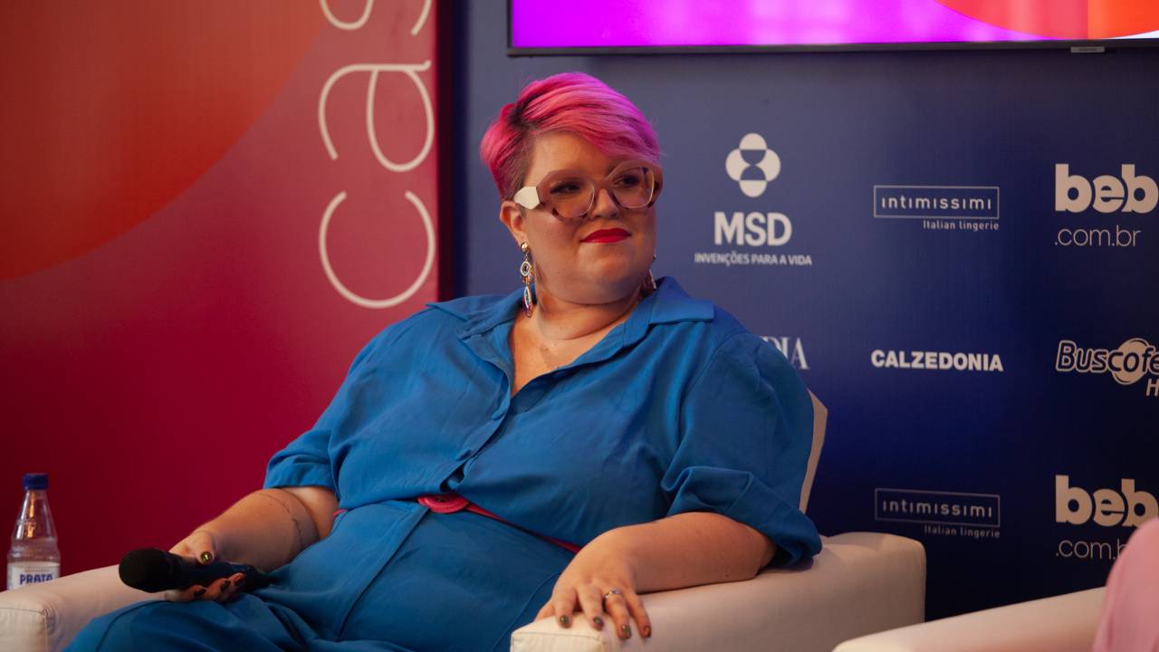 Gabi tem o cabelo curto e rosa, veste um macacão azul vibrante e usa um cinto rosa, do mesmo tom do cabelo. Ela está sentada em uma poltrona, com um microfone na mão
