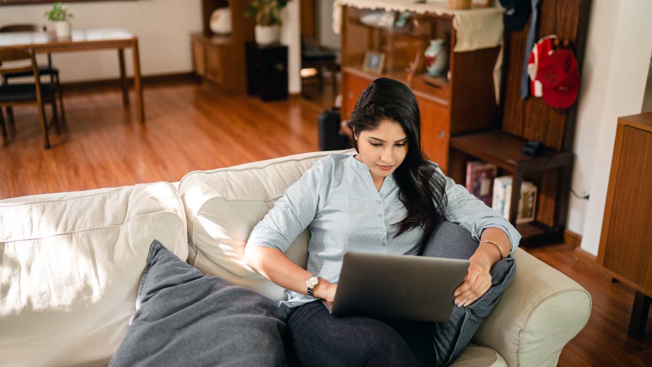 Mulher de cabelo preto liso está com um notebook apoiado no colo, sobre o sofá