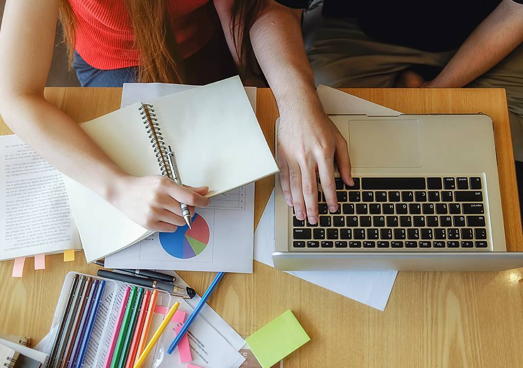 Vista superior de uma mesa de estudos com um notebook, cadernos, folhas e cadernos sobrepostas. Há duas pessoas atrás da mesa com os braços apoiados sobre ela.