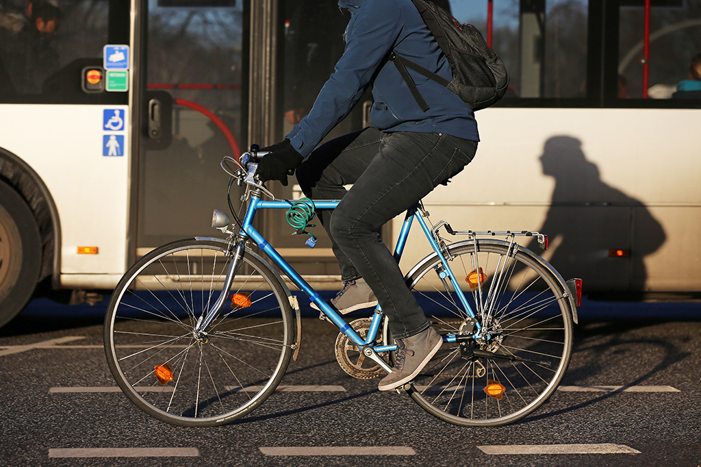 Homem andando em bicicleta. Há um ônibus ao fundo.
