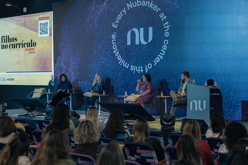 Pessoas sentadas diante de um palco no qual três mulheres e um homem estão sentados.