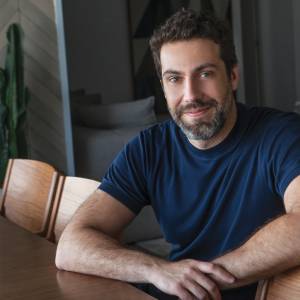 VCRH_94_Líderes_solidão_2 Foto de um homem de barba sorrindo vestindo camiseta azul, sentado a mesa.