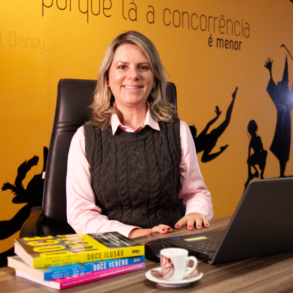 VCRH_96_Assédio_Moral_1 Imagem de uma executiva sorrindo, sentada diante de um notebook, vestindo uma camisa rosa e um colete preto.