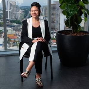 VCRH_97_Diversidade_2 Imagem de uma mulher sorrindo, sentada ao lado de um grande vaso preto. Vestindo um conjunto preto e branco. Ao fundo, através da janela, vê-se a cidade.