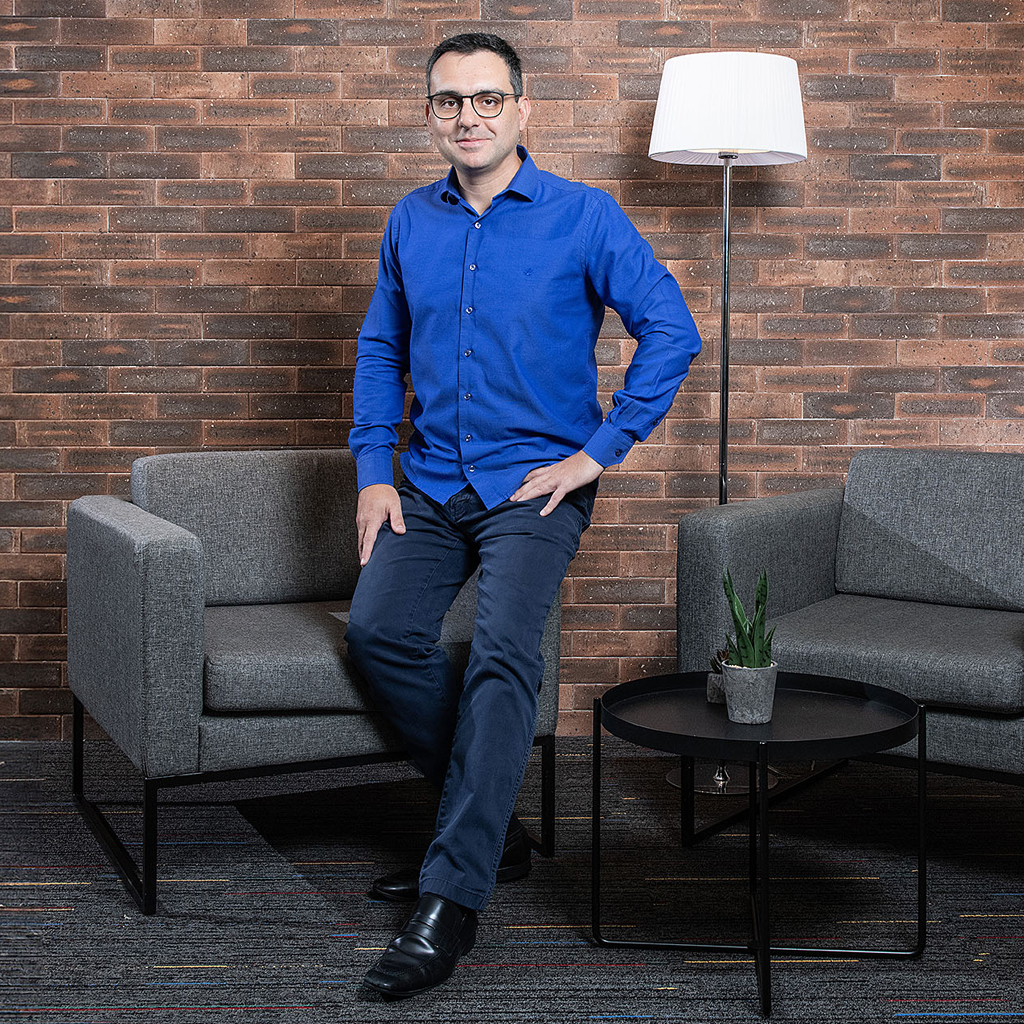 Imagem de um homem sorrindo, vestindo camisa azul, de p&eacute; em frente a um conjunto de sof&aacute;s. Ao fundo, uma parede de tijolos aparentes.