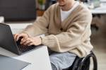 Um homem vestindo um cardigan sentado em uma cadeira de rodas. Ele está digitando no teclado do notebook em cima de uma mesa branca.