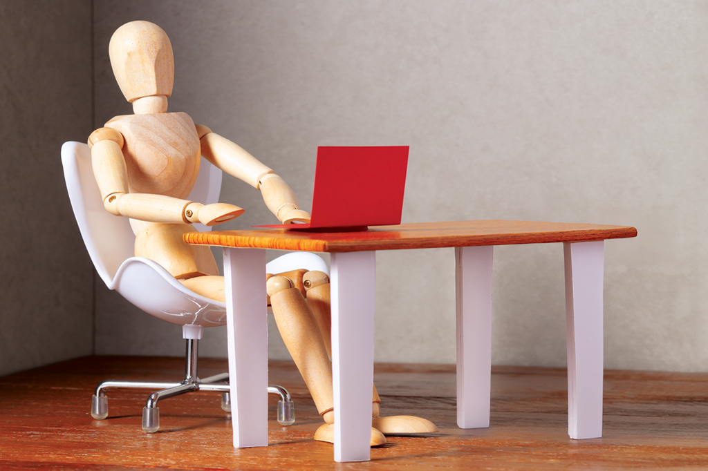 Imagem de um manequim articulado de madeira sentado numa cadeira branca, usando um notebook vermelho que está em cima de uma mesa de tampo de madeira.