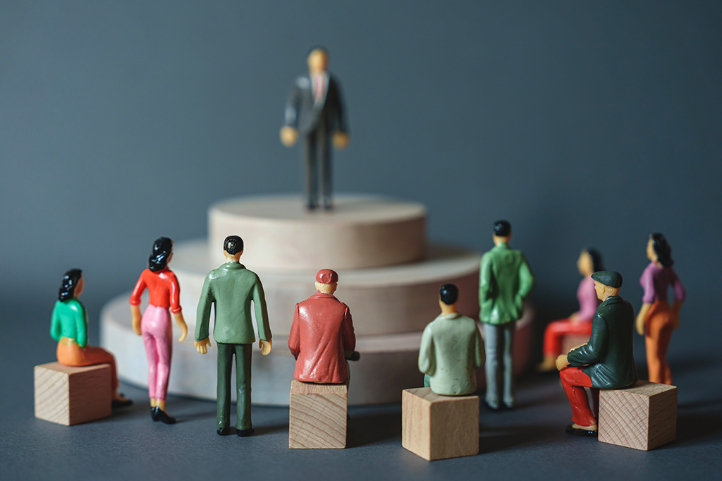 Miniaturas distribuídas em superfície azul, com um homem ao centro em um tablado diante um grupo de pessoas.