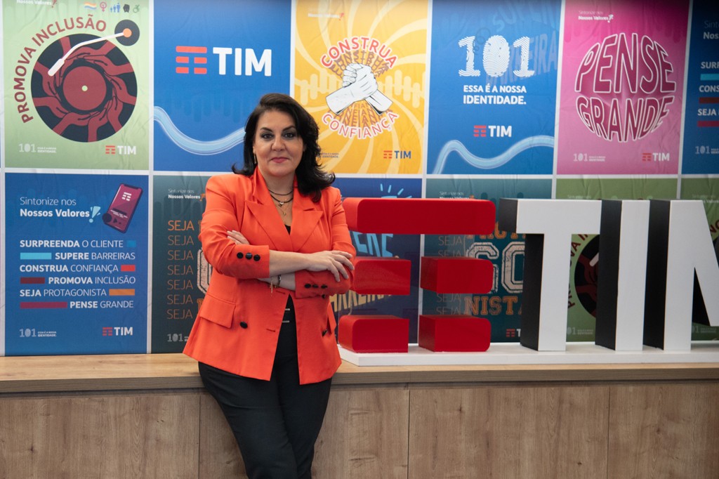 Mulher de meia-idade, cabelos escuros, blazer laranja e calça preta, braços cruzados, sorri levemente. Ela está em um ambiente corporativo com painéis coloridos da TIM ao fundo e um letreiro vermelho e branco com o nome da empresa.