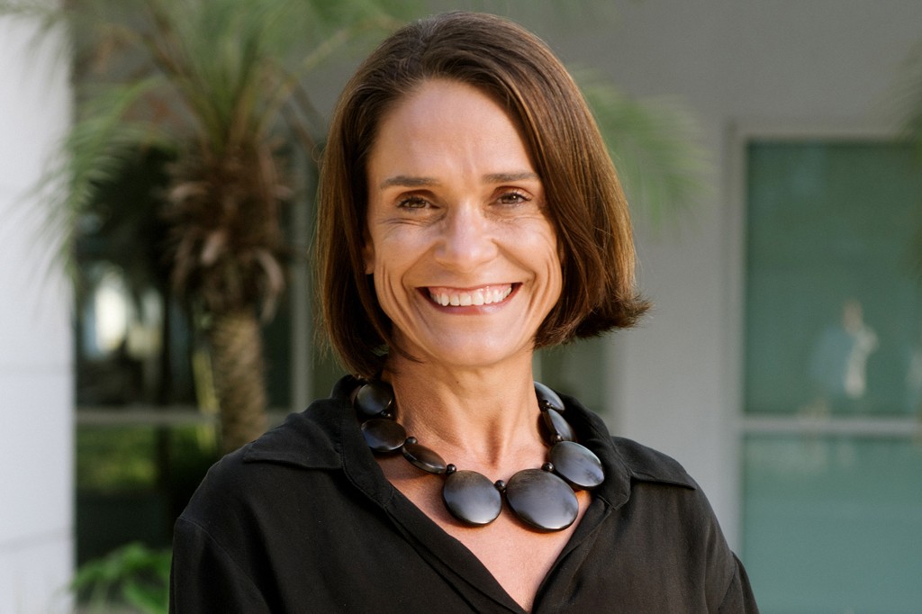 Mulher de pele clara, cabelos castanhos curtos e lisos, sorrindo amplamente, usando uma blusa preta e um colar de contas ovais escuras. Ao fundo, uma palmeira e uma construção clara com janelas.