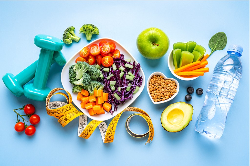 Alimentos saudáveis e itens de exercício sobre um fundo azul claro. Há dois halteres turquesa, uma fita métrica amarela, um prato branco com brócolis, repolho roxo, cenoura picada e tomate cereja. Também aparecem uma maçã verde, um pote com aipo e cenoura em palitos, um pote em formato de coração com lentilhas, mirtilos, meio abacate e uma garrafa de água