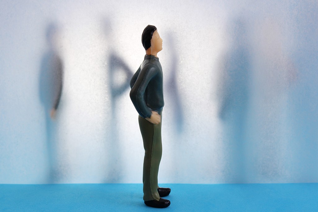 Miniatura de homem de perfil, com suéter azul-escuro e calça verde-oliva, mãos na cintura, olhando para a direita. Ao fundo, sombras de pessoas embaçadas contra uma parede translúcida azul-clara, sobre uma superfície azul-escura