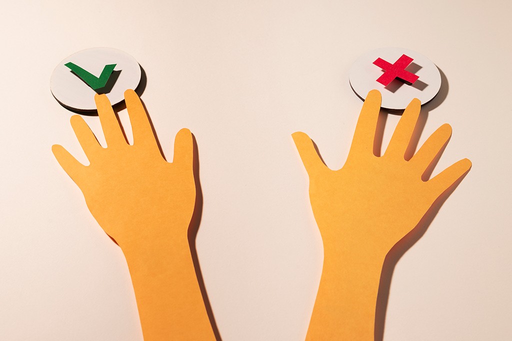 Duas mãos de papel laranja, uma à esquerda com um sinal de certo verde e outra à direita com um X vermelho, sobre um fundo claro